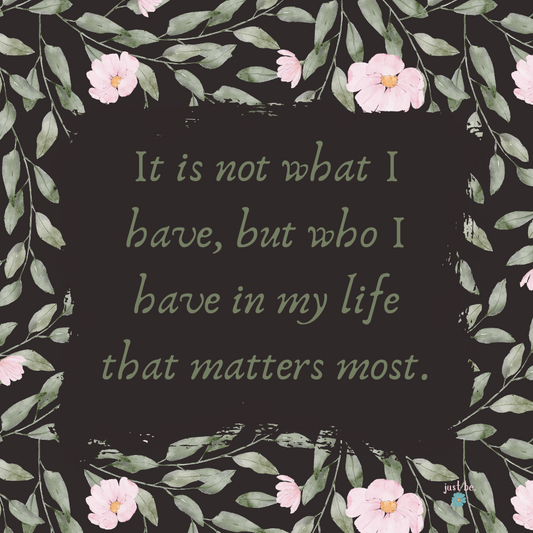 "It is not what I have but who I have in my life that matters most" written in an elegant script on a dark background with soft pink flowers and green leaves surrounding the text.